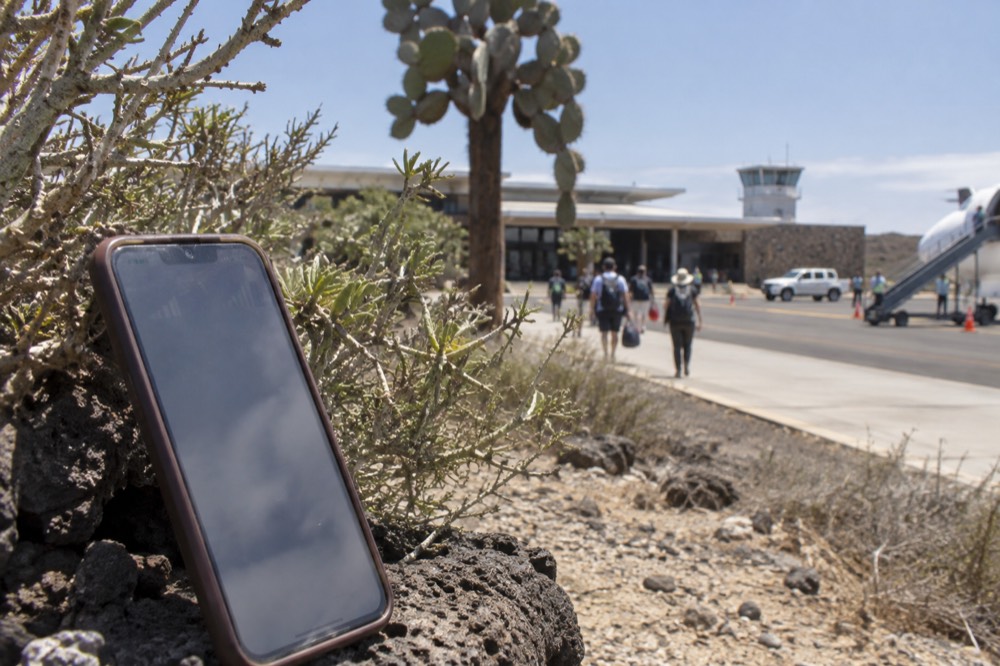 A phone propped against a saline bush at Baltra in the Galapagos, Ecuador, signal bars hunted on a sun-bleached screen, cactus and airport pale behind