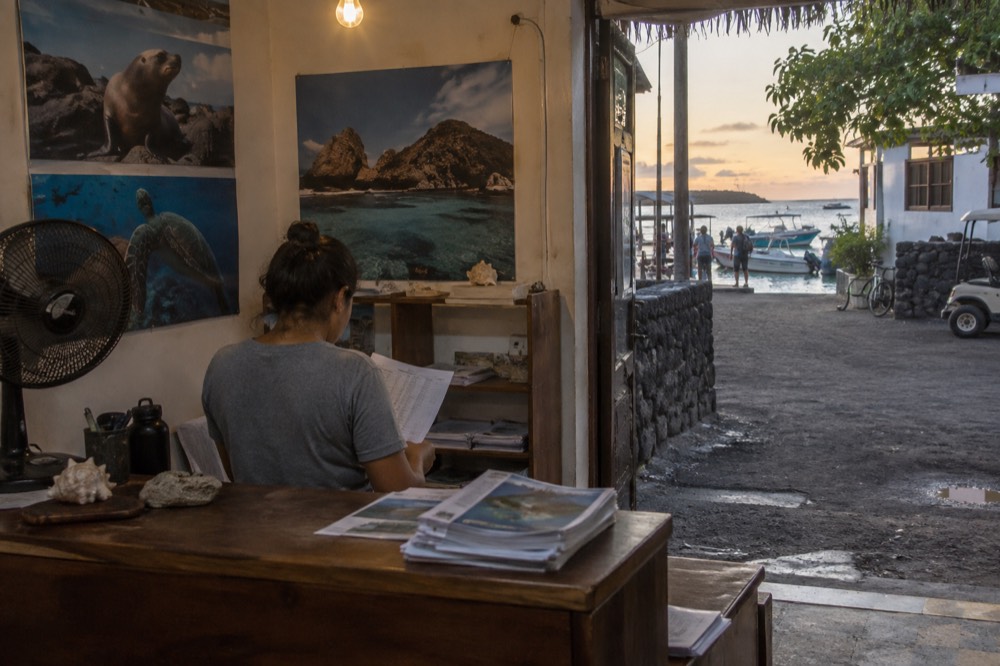 Inside a small tour office on an unpaved street in Puerto Ayora, Galapagos Islands, Ecuador, early evening; a scuffed counter with loose ferry timetables
