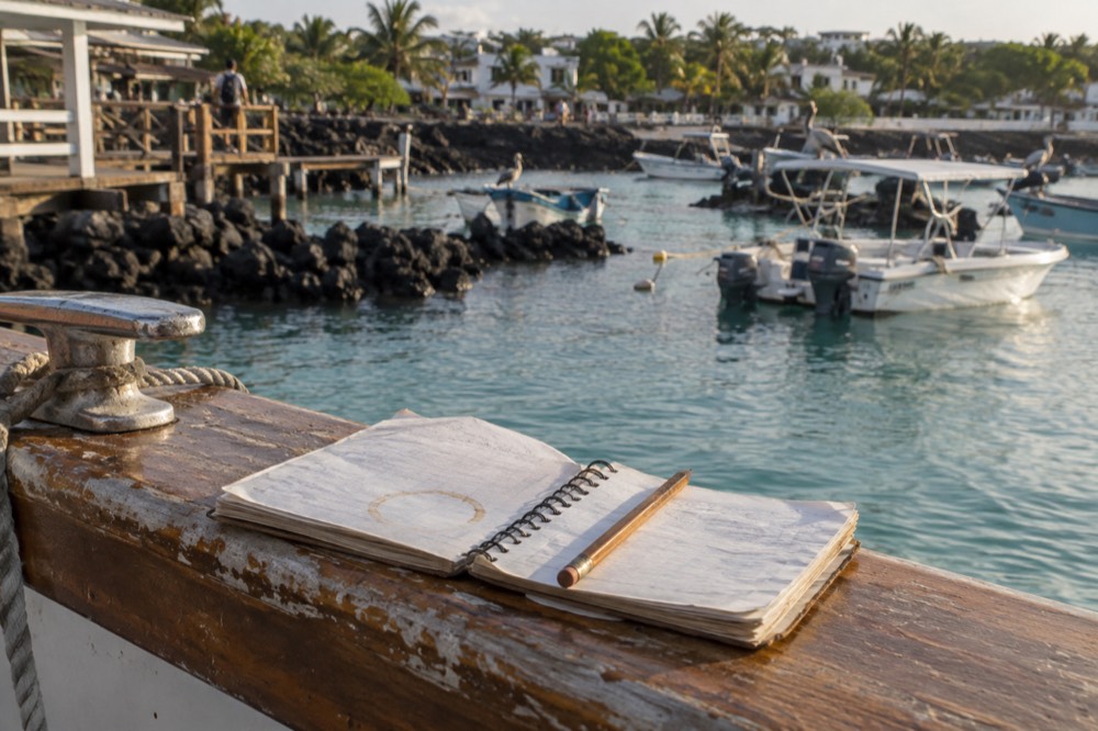 A weathered price-comparison notebook and pencil resting on a salt-stained teak boat rail at Puerto Ayora dock, Galapagos Islands, Ecuador