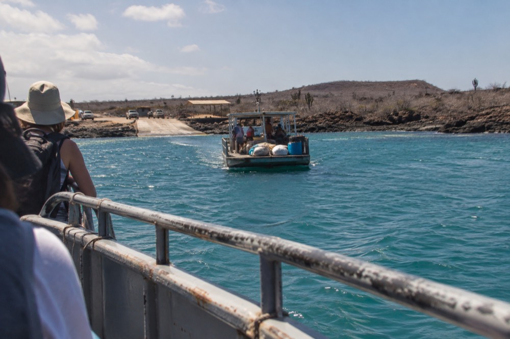 The ferry channel at Baltra Island, Galapagos Islands, Ecuador, mid-morning glare, a small wooden ferry loaded with packs mid-crossing toward Santa Cruz